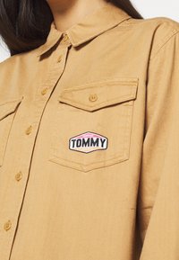 Camicia button-up marrone chiaro realizzata in tessuto con una texture liscia, caratterizzata da due tasche a soffietto e un logo sul petto sinistro.