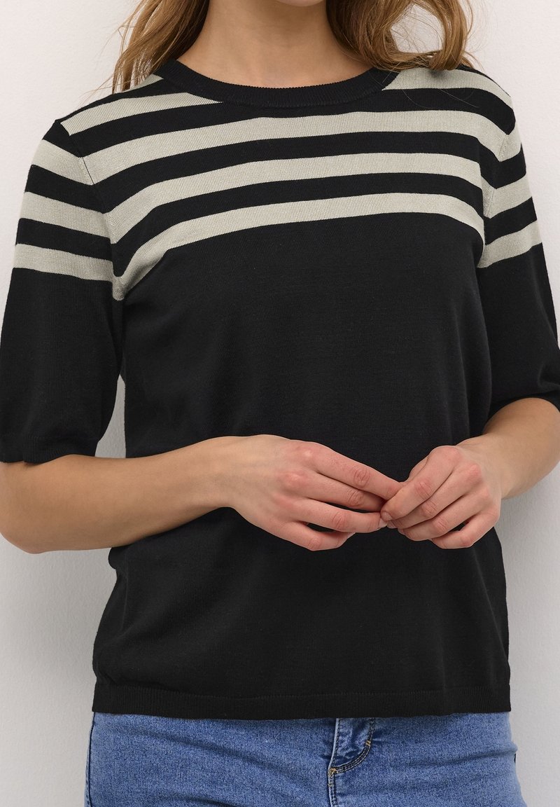 Femme portant un haut noir à manches courtes avec des rayures horizontales beige près de l'encolure, associé à un jean denim bleu, les mains doucement jointes.