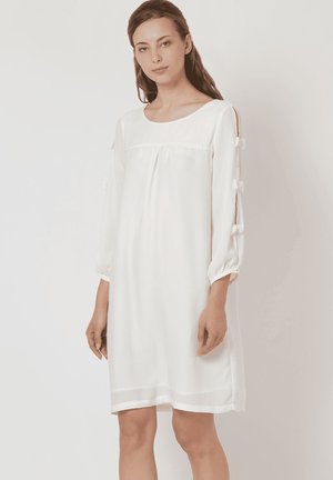 Vestido blanco hecho de tela ligera, con un corte holgado, cuello redondo y mangas abullonadas con acentos de lazo en los laterales.