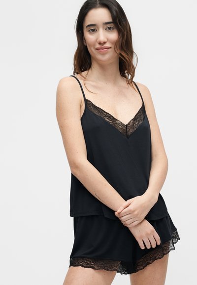 Γυναίκα με μαύρο σετ καμισόλας και σορτς με δαντέλα, στάση με τα χέρια σταυρωμένα, ουδέτερο ανοιχτό γκρι φόντο.