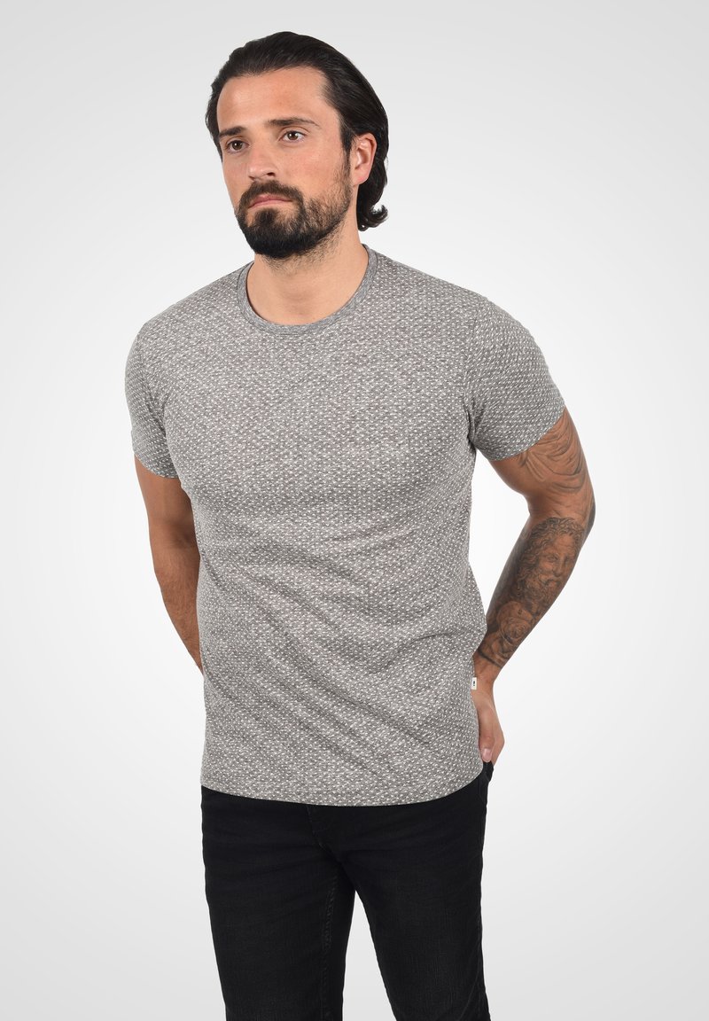 Tricou gri cu mânecă scurtă, având un model texturat. Materialul pare ușor și ușor elastic. Poartă-l cu pantaloni negri.