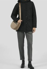 Person som bär en svart knappad kappa, grå rutiga byxor, svarta stövlar och bär en beige axelväska mot en ljusgrå bakgrund.