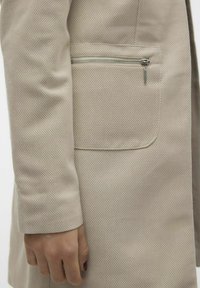 Beige kabát s herringbone texturou, s boční kapsou na zip a přiléhavým střihem se strukturovanými rukávy.