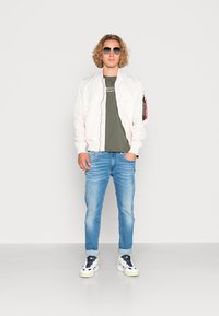 Lysfarvet bomberjakke, olivengrøn t-shirt, lysblå jeans og hvide sneakers med sorte accenter; står foran en ensfarvet baggrund.