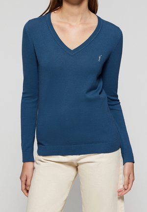 Femme portant un pull bleu uni à manches longues avec col en V et un petit logo brodé sur la poitrine ainsi qu'un pantalon beige.