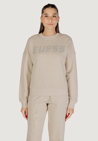 Felpa beige realizzata in morbido tessuto, con scollo rotondo, spalle scese e un logo scintillante "GUESS" sulla parte anteriore.