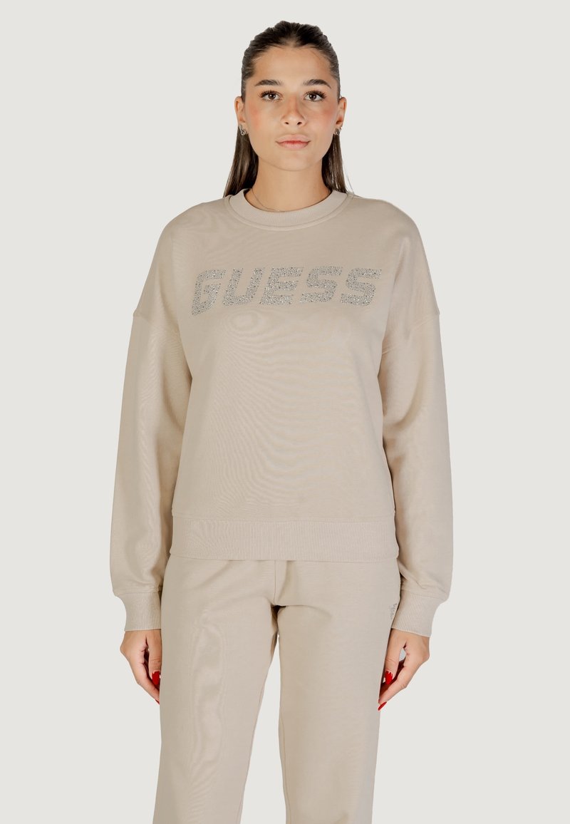 Felpa beige realizzata in morbido tessuto, con scollo rotondo, spalle scese e un logo scintillante "GUESS" sulla parte anteriore.