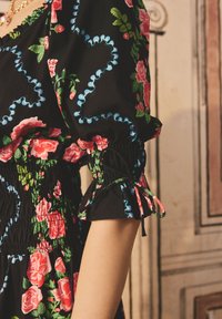 Rochie florală neagră cu trandafiri roșii, frunze verzi și accente albastre. Prezintă mâneci bufante cu manșete plisate și un tors smockat.