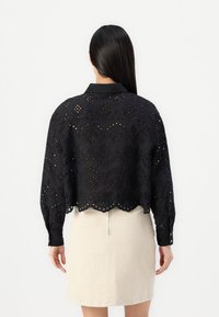 Black embroidered blouse with a cropped silhouette, featuring openwork patterns and scalloped hem; paired with a beige skirt.