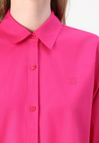 Pink button-up shirt with a collar, featuring a subtle embossed logo. Smooth texture and tailored design. Buttons match the fabric color.