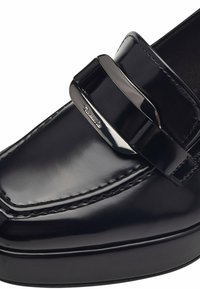 Mocassin en cuir verni noir avec un bout arrondi, présentant une finition brillante et un accent de bande métallique sur le dessus. Détails de couture visibles.