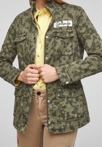 Persoon in een groene camouflagejack met een "Dreamers" patch, een gele overhemd, en beige broek, met de handen gekruist op de rits van het jack.