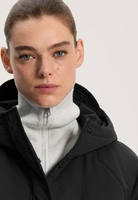 Črn prešit jopič z visokim ovratnikom in sivi rebrasti pulover pod njim; ima zadrgo in gumbasto zapiranje ter teksturirano tkanino.