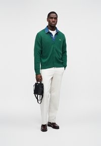 Χακί κασκόλ με V-λαιμό και κουμπιά, φορεμένο πάνω από πουκάμισο τζιν, συνδυασμένο με κρέμες παντελόνια και μαύρες δερμάτινες γόβες. Κρατιέται μια μικρή μαύρη τσάντα.