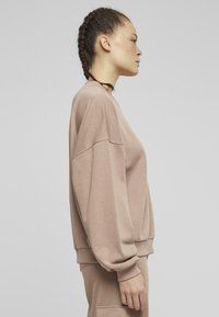 Felpa beige oversize con scollo rotondo, spalle cadute, polsini a coste e una consistenza morbida. Tessuto liscio su uno sfondo semplice.