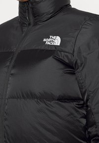 Gros plan d'une doudoune noire avec un col montant, une fermeture éclair à l'avant et le logo blanc "The North Face" sur la poitrine.