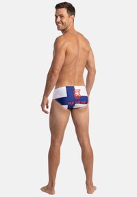 Hymyilevä mies seisoo paljain jaloin, ilman paitaa, valkoiset uimahousut joissa sininen risti, punainen teksti "MR UKKO" ja punainen kasvologo selässä.