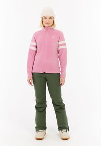 Pullover in pile di colore rosa chiaro con strisce bianche sulle maniche, colletto con zip, abbinato a pantaloni verdi e stivali beige, con orlo regolabile tramite cordoncino.