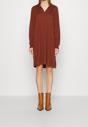 Kobieta w długiej, rdzawej sukience z kołnierzykiem i butach ze skóry w kolorze beżowym sięgających do połowy łydki, stojąca na jednolitym tle.