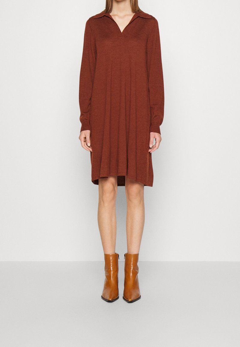 Femme portant une robe à manches longues de couleur rouille avec un col, associée à des bottes en cuir beige mi-mollet, debout devant un fond uni.