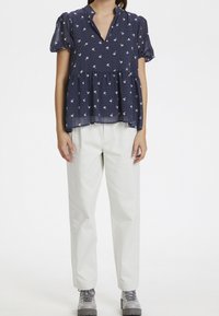 Blusa blu navy a maniche corte con design a peplo, motivo floreale bianco e scollo a v, abbinata a pantaloni bianchi e scarpe grigie.