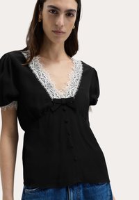 Schwarze Bluse aus Stoff mit einem weißen Spitzen-V-Ausschnitt, Puffärmeln und einem kleinen Schleifenakzent sowie Knopfdetails an der Vorderseite.
