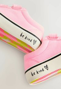 Zapatos de ante rosa con una suela blanca que presenta una inscripción negra de "sé amable" y un patrón de espiga multicolor en la parte inferior.