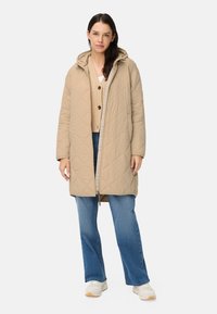 Beiger gesteppter Mantel mit Kapuze, ausgestattet mit einem Frontreißverschluss, Seitentaschen und einem lockeren Schnitt, kombiniert mit einer beigen Strickjacke und blauen Jeans.