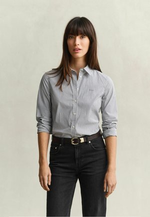 Jeune femme aux cheveux bruns raides, portant une chemise rayée boutonnée et un jean noir, debout devant un fond clair uni.