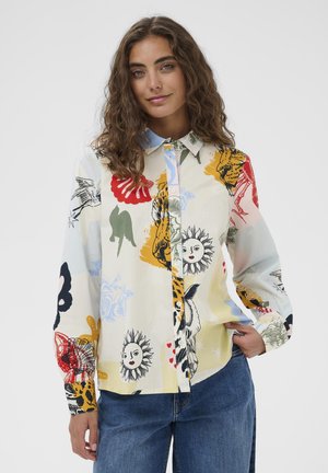 Woman with wavy brown hair wearing multicolored button-up shirt with sun, flower, bird, and abstract prints, paired with blue jeans.