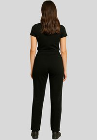 A woman with long hair wears a fitted black top and high-waisted black pants, standing against a neutral gray background.