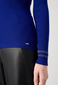 Blå ribbad långärmad topp med guldfärgade ränder på ärmsluten. Tyget ser mjukt och åtsittande ut, kombinerat med svarta skinnbyxor.