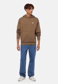 Felpa marrone con tasca frontale e coulisse, abbinata a jeans blu e sneaker bianche, in piedi di fronte a uno sfondo semplice.