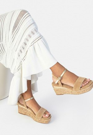 Vrouw draagt beige sleehakken met enkelbandjes en een witte midi-rok met textuur, zittend tegen een witte achtergrond.