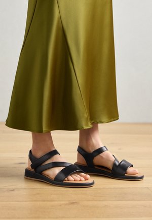 Sandales en cuir noir avec trois sangles horizontales et une bride de maintien au niveau de la cheville, dotées d'une semelle en caoutchouc plate et d'un design minimaliste.