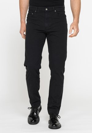 Uomo che indossa pantaloni neri slim fit e scarpe eleganti nere lucide, in piedi su sfondo bianco.