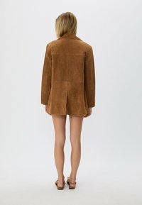 Person med blont hår klädd i en brun mockajacka och bruna sandaler, stående vänd bortåt mot en enkel ljus bakgrund.