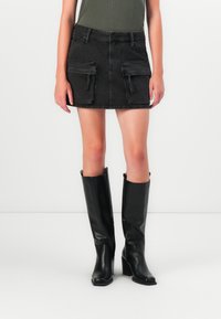 Gonna mini in denim nero con due tasche cargo frontali, dettagli con zip, e vita aderente, abbinata a stivali neri in pelle con tacco al ginocchio.
