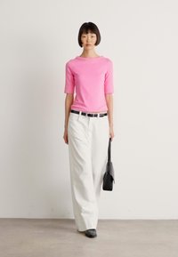 Vrouw in een roze T-shirt met korte mouwen, witte wijde broek, zwarte riem, zwarte schoenen, en met een zwarte handtas, staande tegen een effen achtergrond.