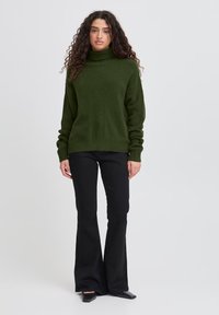 Maglione a collo alto lavorato a maglia verde scuro con polsini e bordo a costine, caratterizzato da un motivo testurizzato che scende verticalmente sulla parte anteriore. Abbinato a jeans neri a zampa.