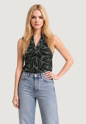 Femme blonde portant un haut noir sans manches à motifs et un jean bleu clair taille haute, debout avec la main sur la hanche devant un fond uni.