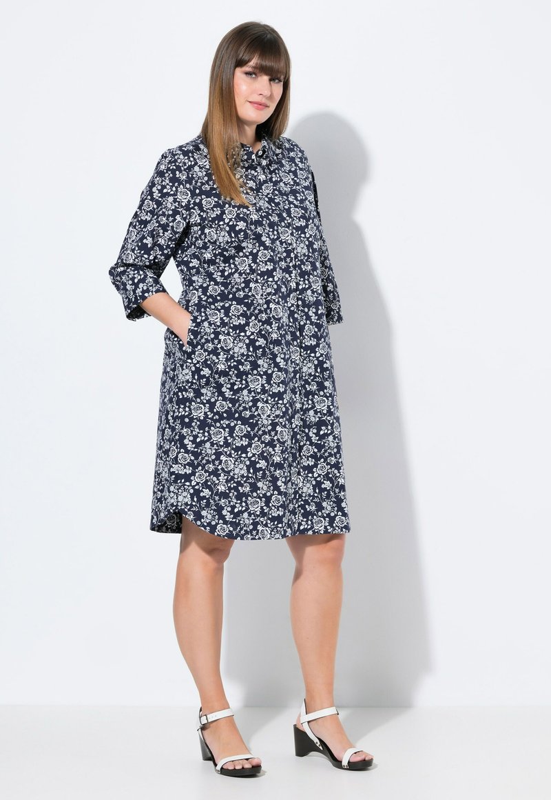 Vestito camicia floreale blu navy con motivi bianchi, con chiusura button-down, maniche a tre quarti e tasche laterali; abbinato a sandali con tacco a blocco bianchi.