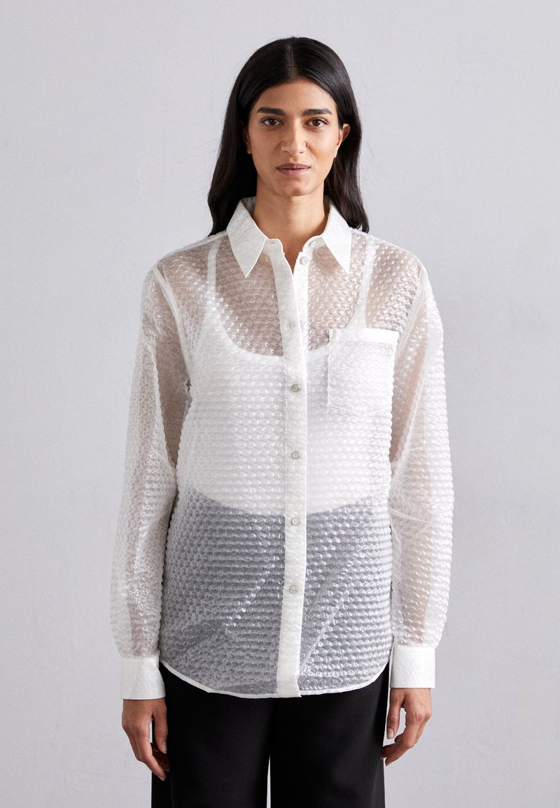 Camicia bianca a maniche lunghe, con motivo, realizzata in un tessuto texturizzato. Presenta una tasca e un fondo grigio a contrasto. Materiale leggero e trasparente.