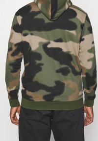 Persona con un hoodie camouflage verde, nero e marrone, indossato con pantaloni scuri, vista da dietro su uno sfondo semplice.