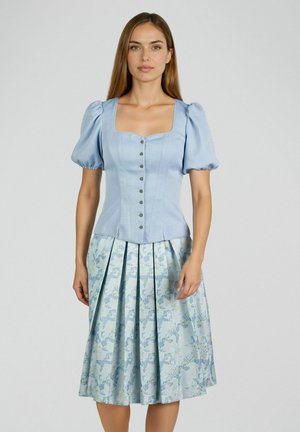 Femme portant un chemisier ajusté bleu clair avec des manches ballon et une jupe midi plissée à fleurs bleues, debout devant un fond uni.