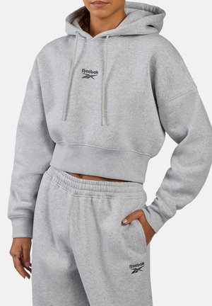 Woman wearing a light gray cropped Reebok hoodie and matching sweatpants with logos on chest and thigh, hand resting in pocket.