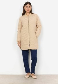 Beige vadderad lång jacka med stående krage, dragkedja framtill och sidofickor, tillsammans med marinblå byxor och beiga ballerinas.