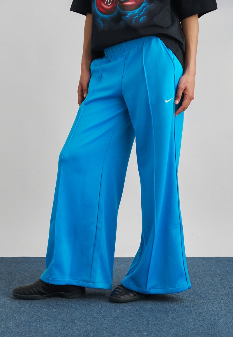 Άτομο που φοράει φαρδιά, φωτεινά μπλε αθλητική φόρμα Nike και μαύρα παπούτσια χωρίς κορδόνια, στέκεται σε μπλε χαλί σε απλό γκρι φόντο.