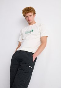 T-shirt blanche avec impression graphique verte, associée à un pantalon noir matelassé avec une poche et un logo. Coupe décontractée, en coton lisse et tissu texturé.