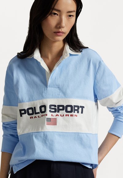 Gaiši zils ilgstoša piedurkņu polo krekls ar baltu horizontālu svītru, kurā ir uzraksts "POLO SPORT" treknās melnās un sarkanās burtiem, kā arī ar amerikāņu karoga akcentu.